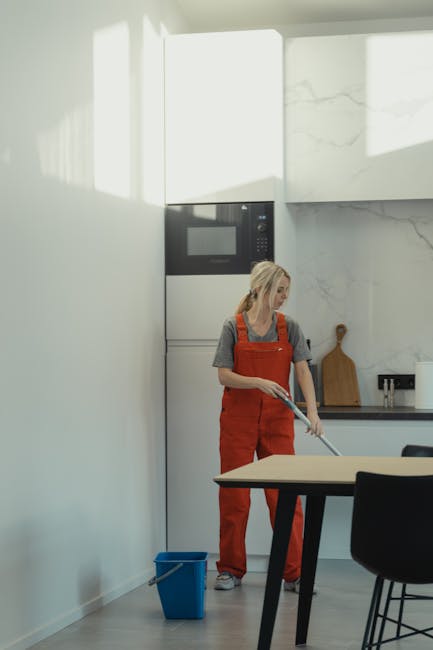 A woman leaning out of a second-floor window with a floral-patterned blanket hanging over the sill. She is wearing a headscarf and appears to be engaged in an outdoor cleaning or dusting task. The win