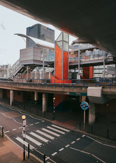 A red and blue DLR train at a station platform with large windows and clean stainless steel doors, set against a backdrop of modern buildings under a cloudy sky with light snowfall. The train appears
